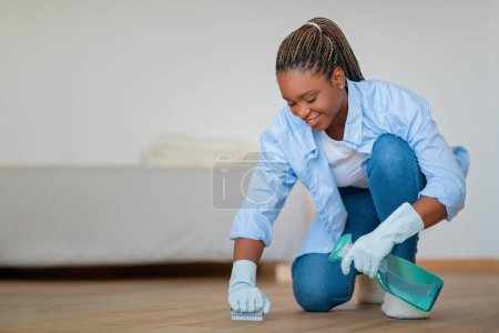 Photo for Cheerful attractive young black woman standing on one knee, wearing rubber gloves, using spray and brush, cleaning wooden floor at apartment and smiling, easy cleaning concept, copy space - Royalty Free Image