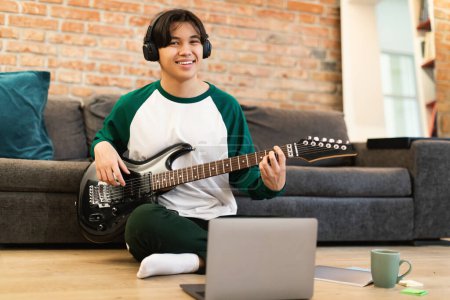 Foto de Japonés adolescente chico jugando música en la guitarra eléctrica con auriculares cerca del ordenador portátil sonriendo a la cámara sentado en el piso en casa. Talentoso joven músico aprendizaje acordes en fin de semana. Enfoque selectivo - Imagen libre de derechos