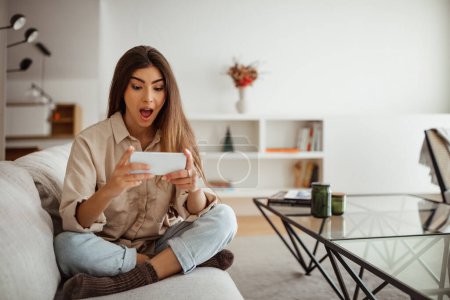 Photo for Cheerful shocked millennial mixed race female with open mouth looks at phone, reads message, plays game, enjoy free time in room interior. Win, app, good news emotions, surprise at home - Royalty Free Image