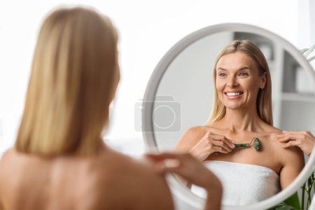 Téléchargez les photos : Traitements pour la peau. Belle femme du Moyen Âge utilisant le rouleau de jade de Greenstone à la maison, heureuse femme mûre faisant le massage de levage de corps avec l'outil de beauté tout en regardant le miroir dans la salle de bain - en image libre de droit