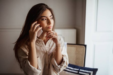 Photo for Thoughtful serious millennial mixed race lady calls by smartphone, talks, thinks, sits on chair, looks at empty space in room interior. Communication remotely, gossip, news at home, choose - Royalty Free Image
