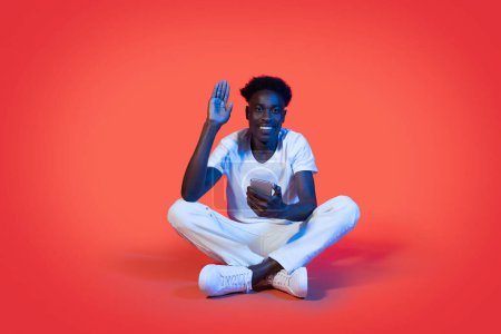 Photo for Cool cheerful young stylish black guy sitting on floor with smartphone in his hand waving and smiling at camera over red studio background in neon light. Telecommunication concept - Royalty Free Image