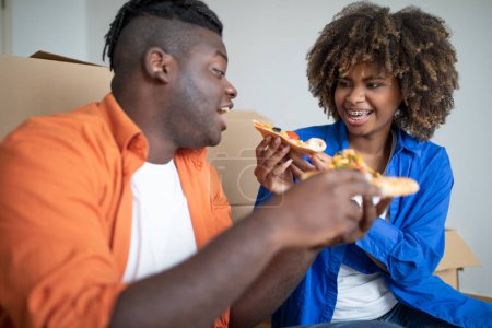 Téléchargez les photos : Joyeux couple noir mangeant de la pizza et s'amusant le jour du déménagement, conjoints afro-américains romantiques se reposant parmi la boxe en carton avec appartenance tout en déménageant à la maison, Femme nourrissant mari, Gros plan - en image libre de droit