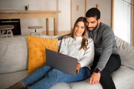 Photo for Smiling international millennial couple sit on sofa, hugging, looking at laptop, have video call in living room interior. Work and rest at home, love and relationship, communication remotely - Royalty Free Image