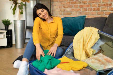 Photo for Happy spanish lady packing clothes in suitcase and talking on smartphone while sitting on floor by couch. Positive female making list notes of things ready to summer travel vacation - Royalty Free Image