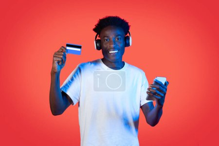 Photo for Cool cheerful handsome young black man hipster with wireless headphones shopping online on red background in neon light, using cell phone and bank card. E-commerce concept - Royalty Free Image