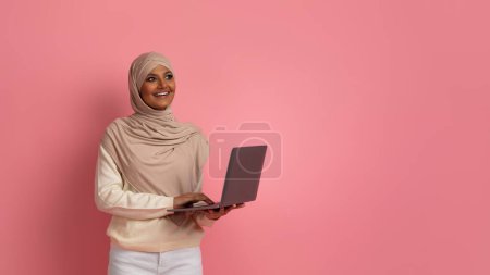 Foto de Mujer musulmana joven pensativa con el ordenador portátil en las manos de pie sobre el fondo del estudio rosa, sonriente dama islámica con pañuelo en la cabeza que sostiene la computadora, disfrutando de oportunidades de trabajo en línea y trabajo remoto - Imagen libre de derechos