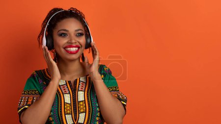 Téléchargez les photos : Joyeux joyeux millénaire dame noire en costume africain traditionnel lumineux en utilisant des écouteurs sans fil, écouter de la musique, en regardant l'espace de copie pour la publicité sur fond de studio orange, panorama - en image libre de droit