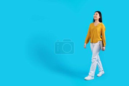 Photo for Happy attractive bright young asian woman in stylish outfit digital nomad with modern laptop walking towards copy space over blue studio background, full length. Freelance, remote job concept - Royalty Free Image