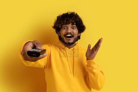 Foto de Feliz joven indio rizado guapo emocional con capucha amarilla que sostiene el control remoto de la TV y gesticulando sobre el fondo amarillo del estudio, disfrutando de programas de televisión, espacio para copiar - Imagen libre de derechos