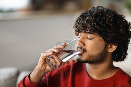 Junger schöner indischer Kerl trinkt zu Hause Wasser aus Glas, Porträt eines Millennial-Hindu-Mannes, der gesundes Mineralgetränk genießt, während er sich auf der Couch im Wohnzimmer entspannt, Nahaufnahme mit Kopierraum