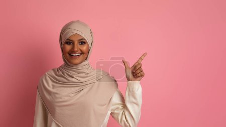 Téléchargez les photos : Belle offre. Souriant femme islamique dans le hijab pointant du doigt à part à l'espace de copie, heureuse musulmane du Moyen-Orient portant foulard indiquant endroit vide sur fond rose pour la conception de la publicité - en image libre de droit