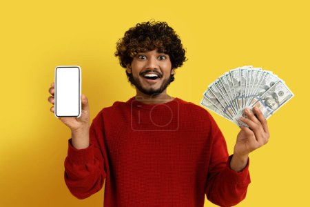Photo for Online loan, credit. Happy handsome young indian man in red sweater showing modern cell phone with white blank screen and cash dollar banknotes, yellow studio background, mockup - Royalty Free Image