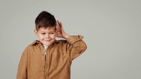 Photo for Smiling interested inquisitive little european boy presses hand to ear listens good news isolated on gray studio background. Pupil emotions, gossip, fun, study and education, ad and offer - Royalty Free Image