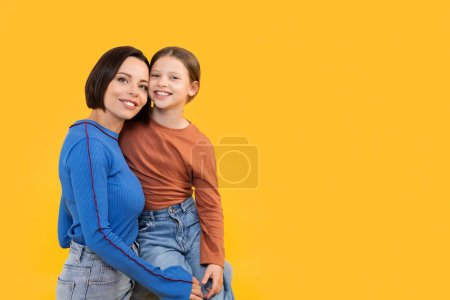 Foto de Retrato de feliz madre joven y linda hijita abrazando mientras posan aislados sobre fondo amarillo, mamá cariñosa y preadolescente niña que se une y sonríe a la cámara, espacio de copia - Imagen libre de derechos