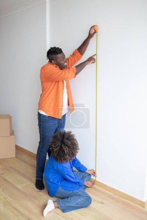 Photo for Happy Young Black Couple Doing Home Repairing, Measuring Wall With Tape Ruler, Cheerful African American Spouses Checking Measurements In Living Room After Moving To New Apartment, Vertical Shot - Royalty Free Image