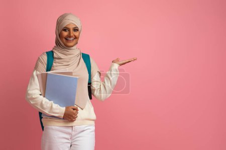 Téléchargez les photos : Étudiante musulmane heureuse dans le hijab pointant vers l'espace de copie avec sa paume, femme arabe souriante portant le foulard et le sac à dos montrant l'objet invisible tout en se tenant debout sur fond rose - en image libre de droit
