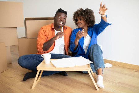 Foto de Cónyuges negros felices discutiendo diseño en nuevo café plano y bebiendo, alegre pareja afroamericana sentada en el piso entre cajas de cartón, lista de necesidades de maling después de mudarse a casa - Imagen libre de derechos