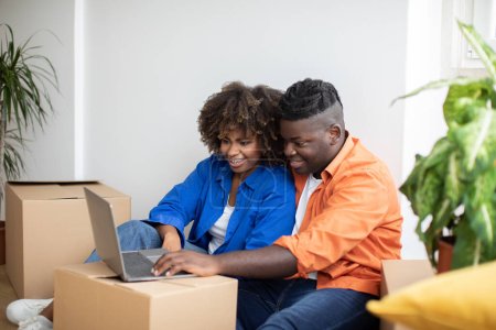 Photo for Moving Concept. Happy Black Couple Using Laptop While Sitting Among Cardboard Boxes In Their New Home, African American Spouses Shopping Online On Computer After Relocation To Own Flat, Copy Space - Royalty Free Image