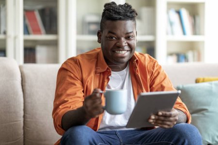 Téléchargez les photos : Positif heureux jeune beau mec afro-américain dans la détente occasionnelle à la maison, assis sur le canapé dans le salon confortable, tenant tablette numérique et tasse à café, espace de copie. Divertissement domestique - en image libre de droit