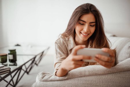 Photo for Glad happy millennial mixed race lady looking at phone, reading message, chatting in social networks, enjoy free time in room interior, free space. Communication remotely, app, good news at home - Royalty Free Image