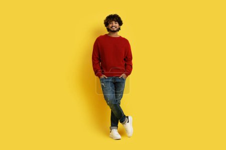 Photo for Happy handsome millennial indian guy in stylish casual outfit with hands in pockets smiling at camera, posing alone on yellow studio background, full length shot with copy space - Royalty Free Image