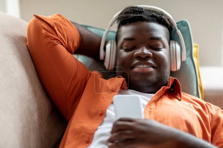 Téléchargez les photos : Relaxé jeune homme afro-américain paisible dans occasionnel couché sur le canapé, en utilisant des écouteurs sans fil modernes et smartphone, écouter de la musique à la maison, profiter d'un son stéréo, copier l'espace - en image libre de droit