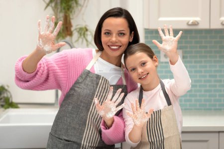 Photo for Happy european millennial female and little girl in aprons cook cookies, show hands with flour, have fun in kitchen interior. Prepare homemade food, hobby in free time together at home - Royalty Free Image