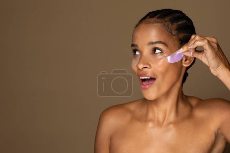Téléchargez les photos : Femme noire excitée d'âge moyen décollant sous les patchs de collagène oculaire, nourrissant la peau douce avec des tampons de gel, espace de copie. Traitement du visage et effet anti-rides - en image libre de droit