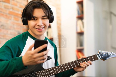 Téléchargez les photos : Asiatique adolescent garçon apprendre à jouer de la guitare électrique en utilisant l'application Smartphone port casque assis à la maison le week-end. Technologie et concept de gadgets musicaux. Focus sélectif - en image libre de droit