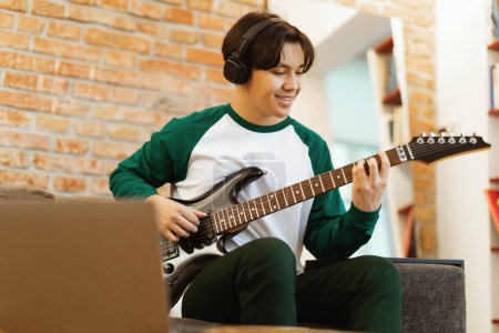 Foto de Happy Asian Teen Guy tocando la guitarra eléctrica y usando la grabación de computadoras portátiles Nueva canción sentada en casa, usando auriculares inalámbricos. Instrumentos musicales y concepto de hobby. Enfoque selectivo - Imagen libre de derechos