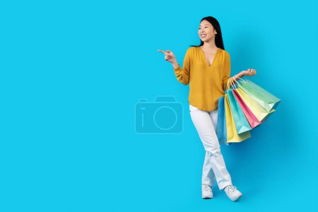 Smiling happy attractive long-haired young korean lady in casual shopaholic showing free space for advertisement over blue studio background, female carrying colorful shopping bags, full length