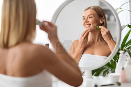 Téléchargez les photos : Belle femme d'âge moyen utilisant le grattoir de jade gua sha à la maison, belle femme mature faisant massage lifting du visage tout en se tenant près du miroir dans la salle de bain, souriant à son reflet, foyer sélectif - en image libre de droit
