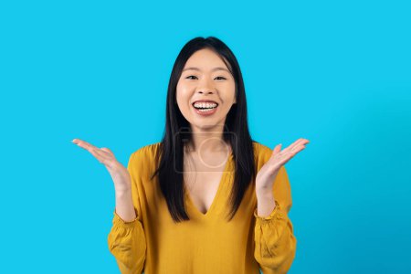 Téléchargez les photos : Émotionnel heureux jeune jolie asiatique femme en jaune geste et rire sur fond bleu studio, femelle montrant l'excitation. Concept d'émotions et de gestes humains - en image libre de droit