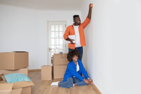 Photo for Young Black Spouses Using Tape Ruler, Taking Measurements In Living Room Together, Happy African American Couple Measuring White Wall, Planning Home Repairing After Moving To New Apartment - Royalty Free Image