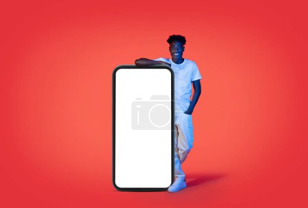 Téléchargez les photos : Cool gai élégant millénariste afro-américain gars en tenue blanche penché sur un grand téléphone portable avec écran blanc blanc maquette espace libre pour l'annonce, montrant belle offre, fond rouge avec néon lumière - en image libre de droit