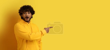 Photo for Amazed emotional excited young curly indian guy in yellow hoodie pointing at copy space for shocking advertisement, isolated on yellow studio background, web-banner, panorama - Royalty Free Image