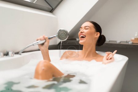 Cheerful Lady Having Fun Taking Bath And Singing Holding Shower Head Like Microphone Sitting In Water With Foam Bubbles In Modern Bathroom At Home. Beauty And Relaxation Leisure