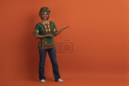 Foto de Mira esto. Sonriente alegre hermosa mujer afroamericana milenaria en traje africano tradicional que muestra el espacio vacío para la publicidad, fondo de estudio naranja, longitud completa - Imagen libre de derechos