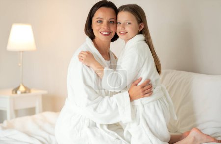 Photo for Beauty care at home together. Glad caucasian teen girl hugging young lady in bathrobe sit on white soft bed, enjoy spare time in bedroom interior. Love, fun, relationship mom and daughter - Royalty Free Image