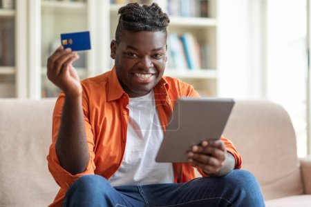 Foto de Feliz chico negro guapo alegre en el cliente casual sentado en el sofá en casa, mostrando tableta digital moderna y tarjeta bancaria azul de plástico, recomendando la banca en línea, compras, espacio de copia - Imagen libre de derechos