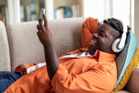 Téléchargez les photos : Souriant chubby millennial black guy dans casual couché sur le canapé avec téléphone cellulaire et écouteurs sans fil, regarder du contenu sur Internet, profiter de l'application divertissante agréable, espace de copie, vue latérale - en image libre de droit