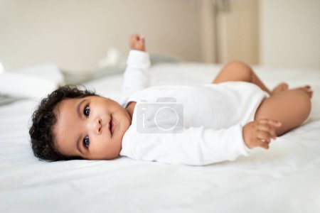 Photo for Cute little african american baby lies on bed, rest and relax, enjoy free time and good morning in white bedroom interior, free space. Child care, childhood, linen for children ad and offer - Royalty Free Image