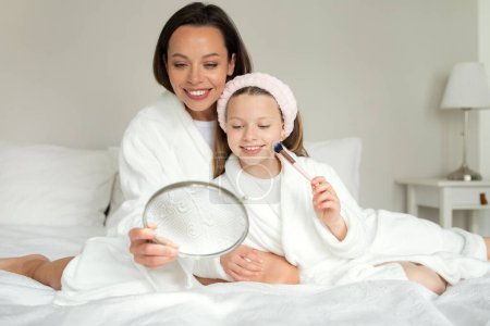 Téléchargez les photos : Joyeux jeune dame caucasienne et petite fille en peignoir s'asseoir sur un lit blanc doux, faire du maquillage, regarder dans le miroir dans l'intérieur de la chambre. Mère fille profiter de temps libre et de soins de beauté à la maison - en image libre de droit