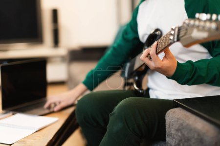Foto de Recortado tiro de chico joven tocando guitarra eléctrica y navegar por Internet en el ordenador portátil para acordes de canción sentado en la sala de estar moderna en casa. Enfoque selectivo en la mano de los músicos - Imagen libre de derechos