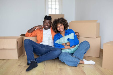 Photo for Moving Day. Happy black couple sitting on floor among cardboard boxes in new home, cheerful african american spouses celebrating relocation, looking and smiling at camera, woman embracing pillow - Royalty Free Image