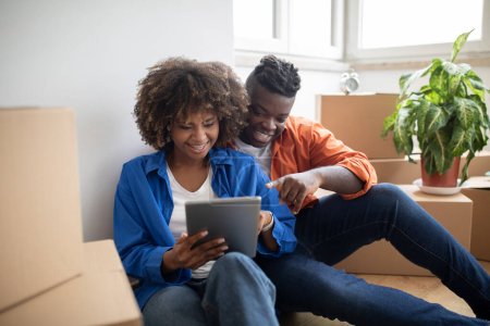 Foto de Feliz pareja negra usando tableta digital mientras está sentada en el piso en un nuevo apartamento entre cajas de cartón con pertenencias después de mudarse, sonriendo cónyuges afroamericanos eligiendo muebles en línea - Imagen libre de derechos