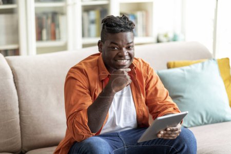Foto de Alegre positivo guapo joven afroamericano chico en casual sentado en el sofá en la sala de estar, utilizando tableta digital, relajarse en fin de semana en casa, espacio para copiar. Aparato moderno, tecnologías - Imagen libre de derechos