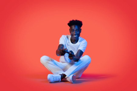 Téléchargez les photos : Joyeux jeune homme noir heureux en tenue décontractée blanche assis sur le sol sur fond de studio rouge au néon, jouant à des jeux vidéo, tenant joystick et souriant, espace de copie - en image libre de droit