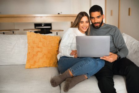 Photo for Glad international millennial family sit on sofa, hugging, watch video on laptop, have online call in living room interior. Communication, rest at home, love and relationship - Royalty Free Image